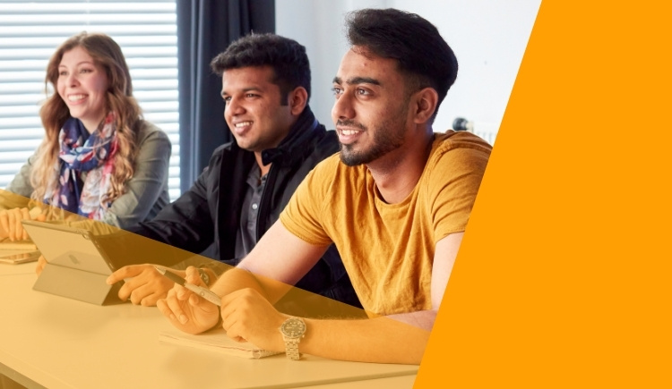 Mae tri o bobl yn eistedd wrth fwrdd, yn edrych ymlaen ac yn gwenu. Mae un person yn dal tabled. Mae'n ymddangos eu bod mewn cyfarfod neu ddosbarth.